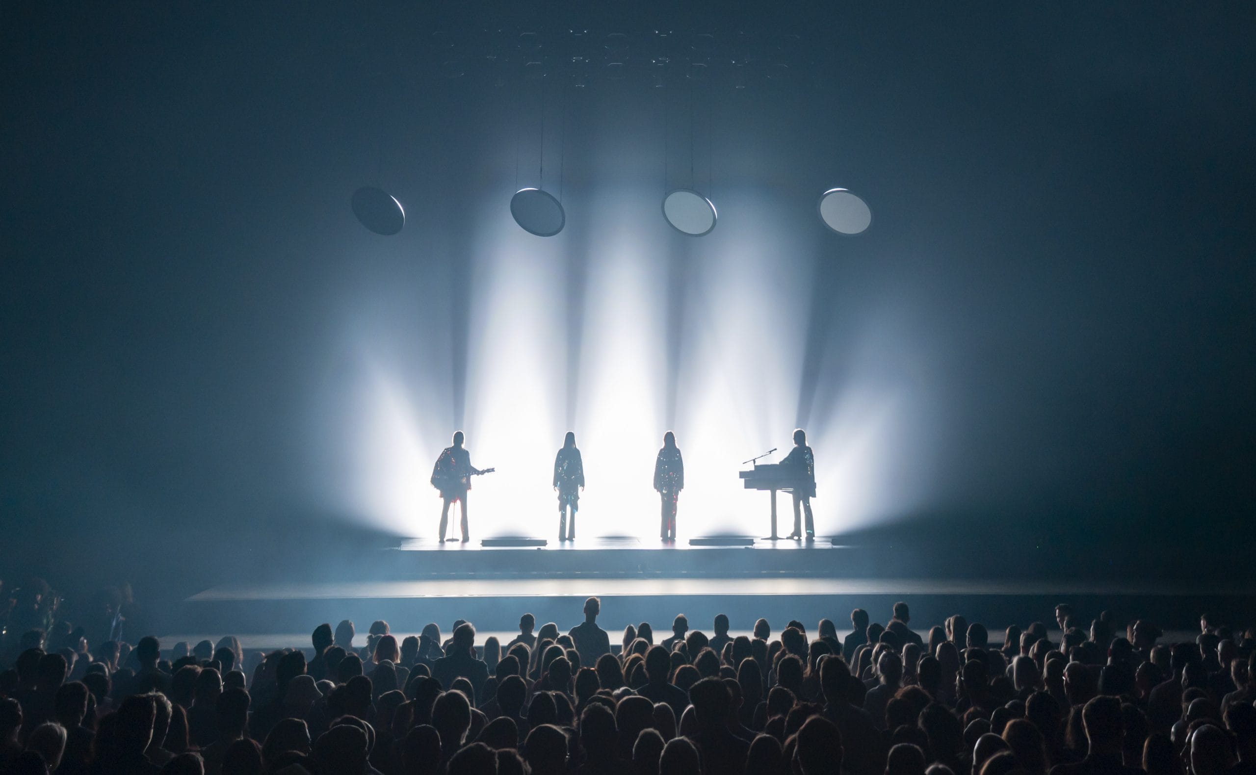 Silhouetted band performs on stage with bright lights.