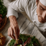 Chef garnishing salad with vegetables and bread nearby.