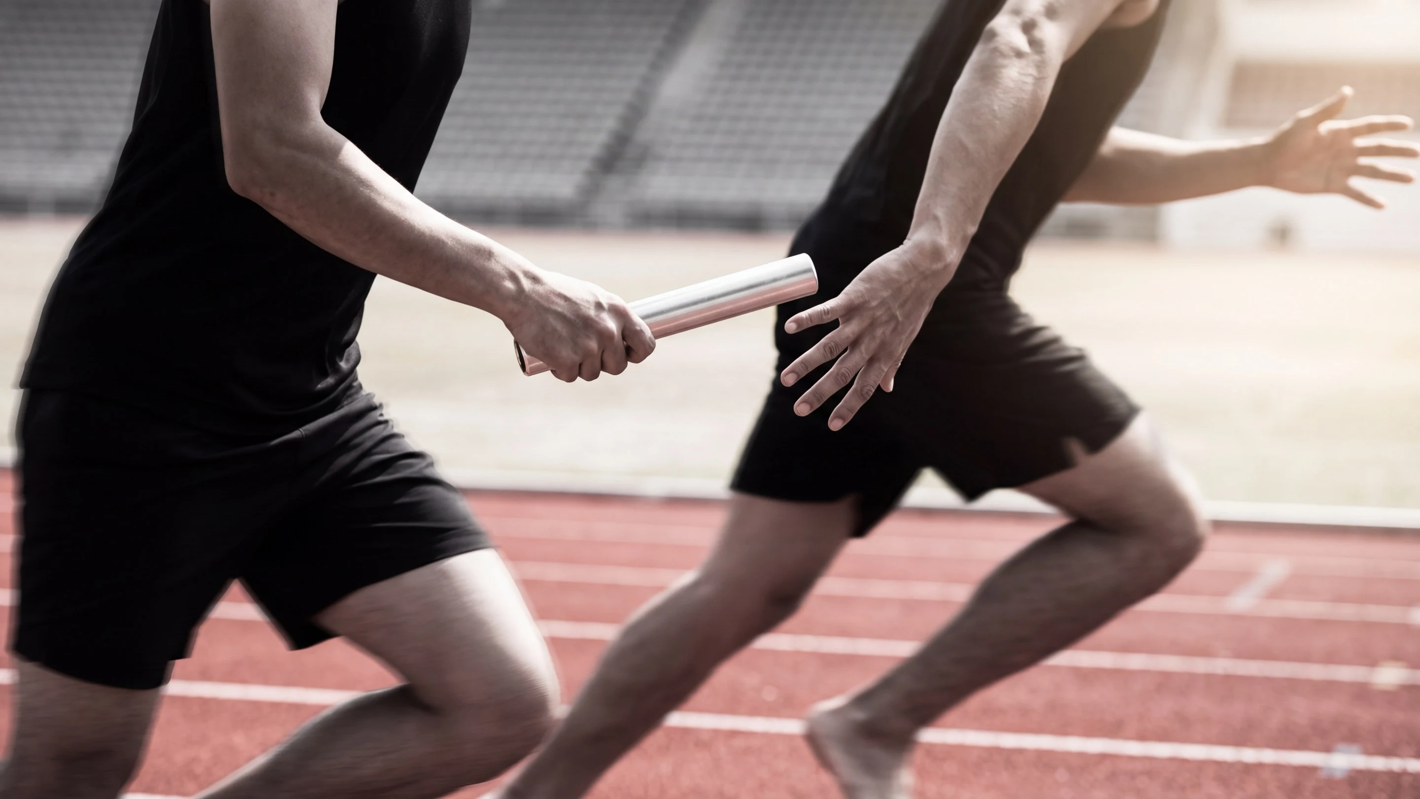 Two runners at full speed, one placing a silver baton into the other’s open hand